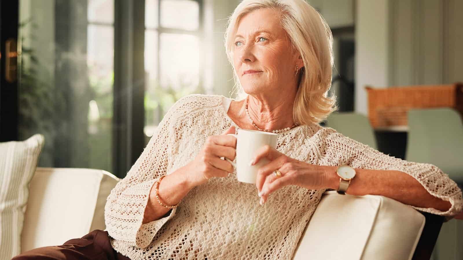 An older woman with gray hair sits on a white sofa, holding a white mug and looking thoughtfully into the distance. Sunlight streams in from behind, creating a warm, peaceful atmosphere.