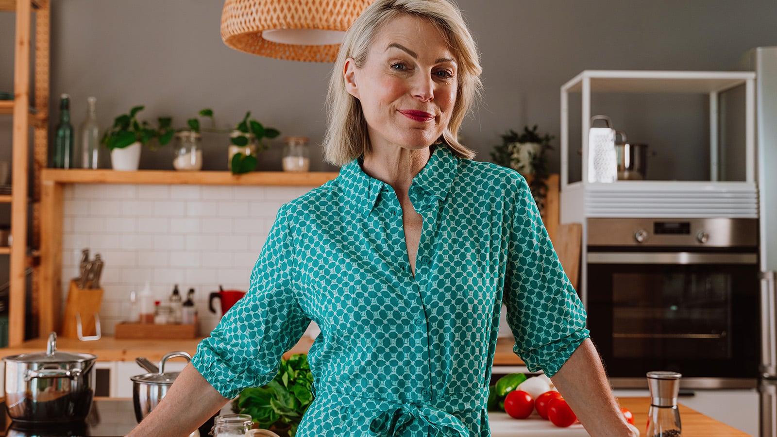 A smiling woman with short blonde hair wearing a teal patterned dress stands in a modern kitchen with wooden shelves, green plants, and fresh vegetables on the counter.