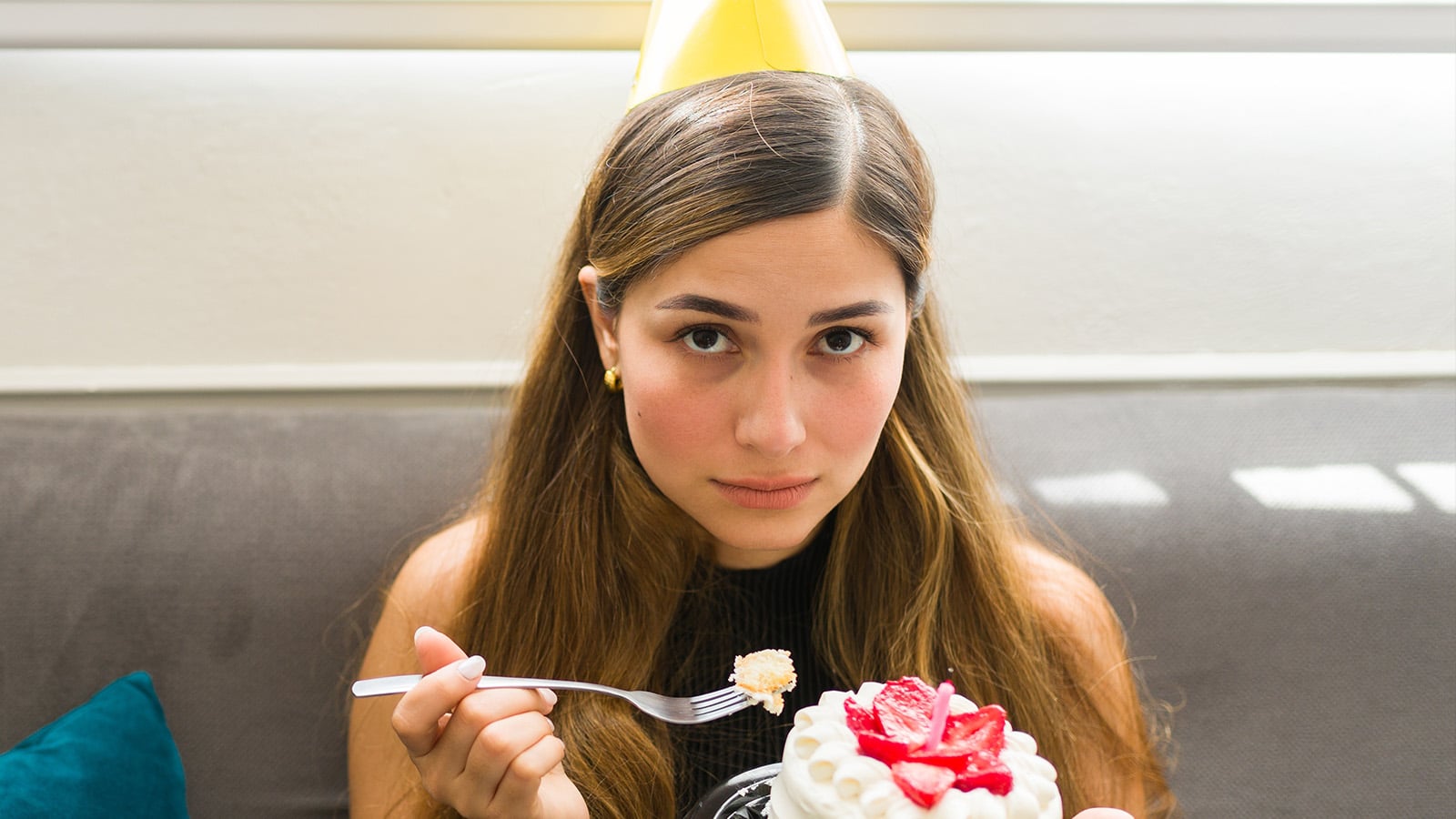 Woman eating birthday cake and wearing birthday hat alone, looking neutral or indifferent