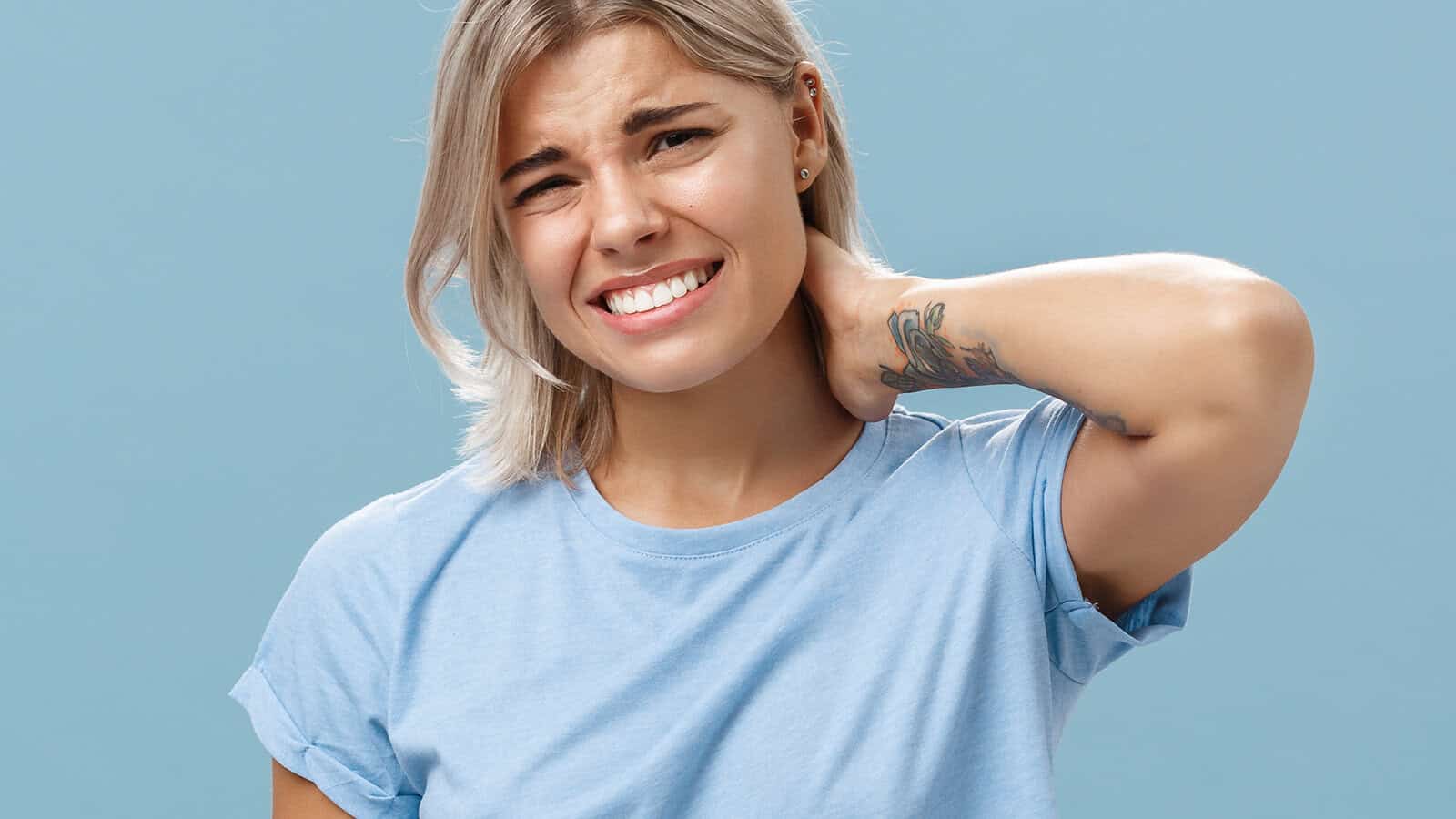 A young woman with short blonde hair wearing a light blue t-shirt looks uncomfortable and holds her neck in pain, grimacing against a plain blue background.