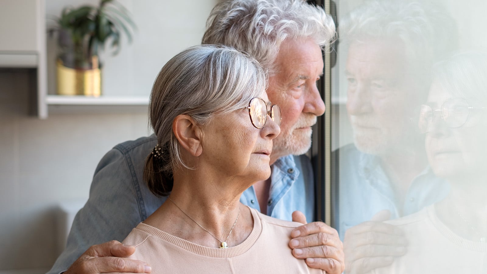 Pensive elderly mature senior couple embracing looking in distance out of window, thinking of personal problems. Old bearded male hugging sad wife close to him, copy space