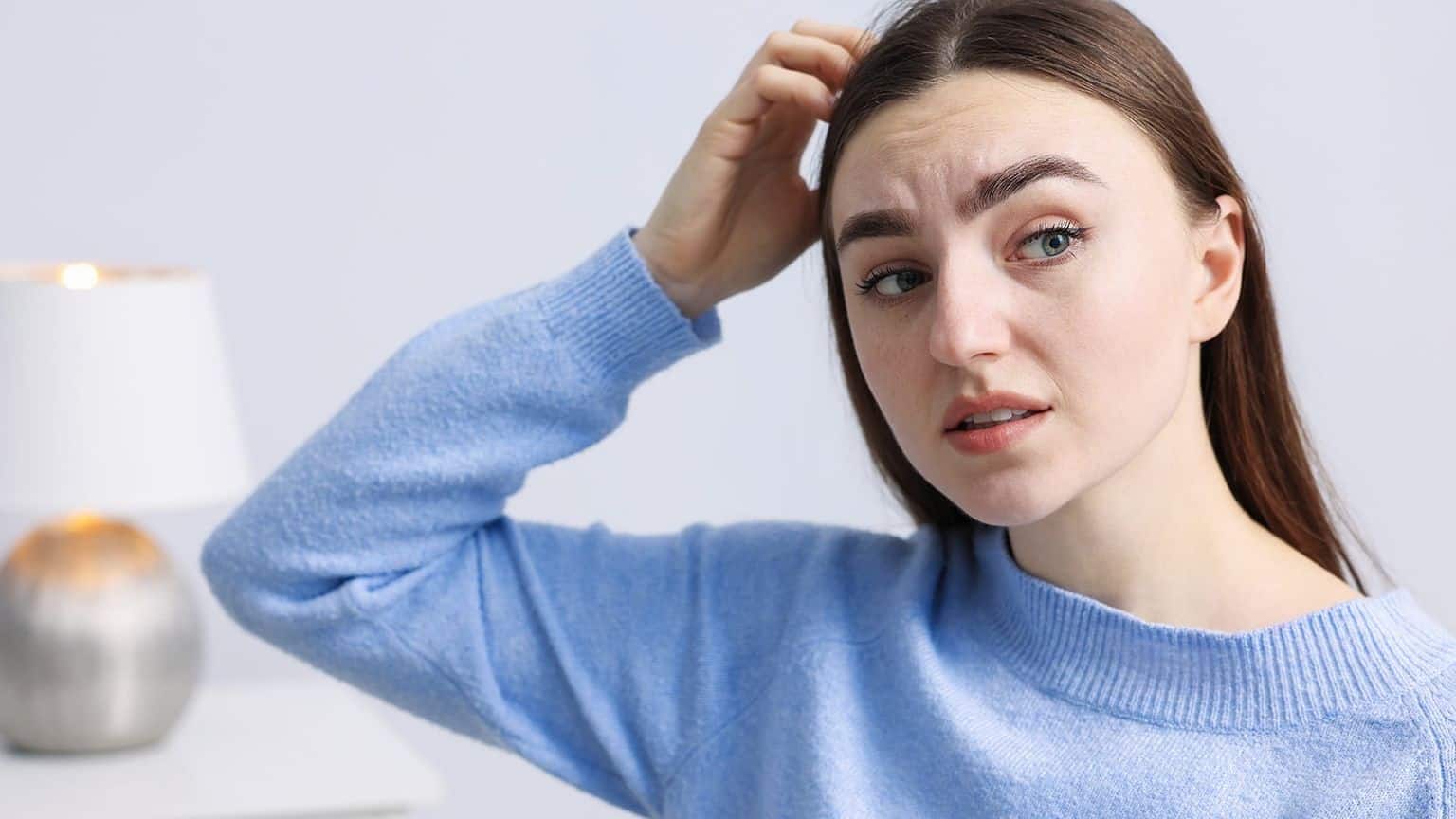 A young woman with long brown hair and a blue sweater touches her scalp while looking concerned, perhaps struggling to remember names during a social interaction. A lamp is blurred in the background.