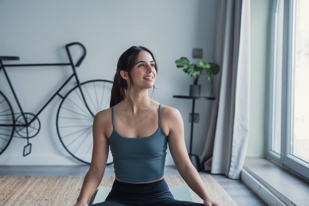 A young woman in workout clothes sits cross-legged on a mat indoors, smiling and looking to the side. Behind her, a decorative bicycle wall piece and potted plant add calm to the space—a serene setting for reflecting on psychology and self-growth.