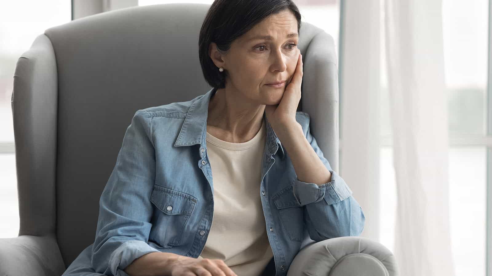 A woman with short dark hair sits in a gray armchair, resting her head on her hand and looking thoughtful or concerned. She wears a light denim shirt over a white top. Light streams in from a window behind her.