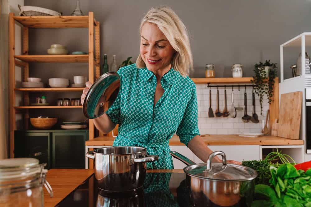 A smiling woman in a teal patterned dress lifts the lid off a pot on the stove, embodying life satisfaction often seen in contented people in their 60s, surrounded by fresh vegetables and kitchenware in her modern, cozy kitchen.