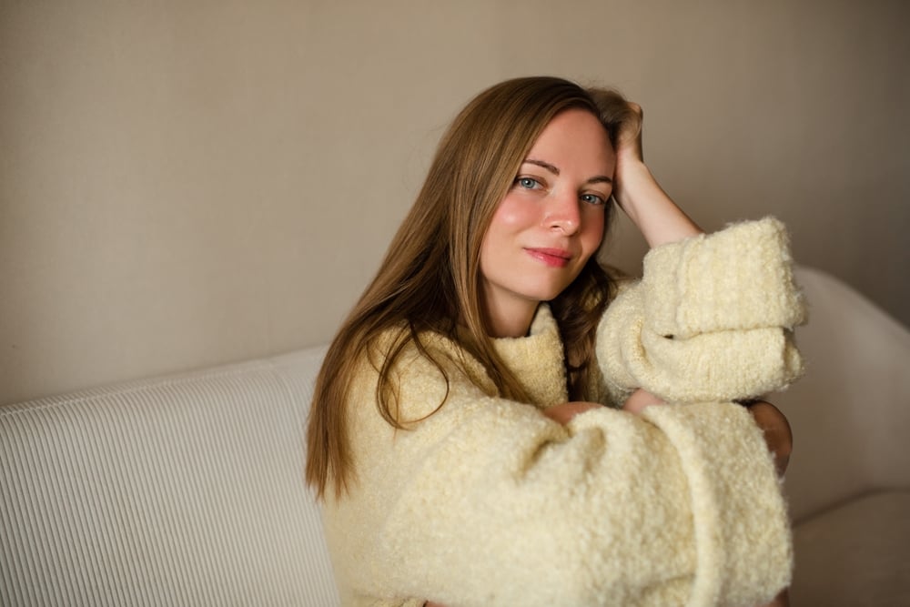 A woman with long brown hair, wearing a cream-colored fuzzy sweater, sits on a light sofa with her head resting on her hand, looking at the camera with a soft smile, reflecting an air of independence and quiet confidence.