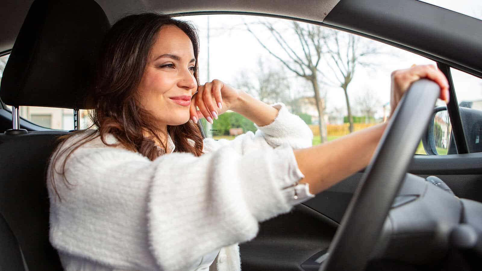 A woman with confident personality traits sits in the driver’s seat of a car, smiling and looking out the window, one hand driving. She wears a white sweater, with trees and houses visible outside.