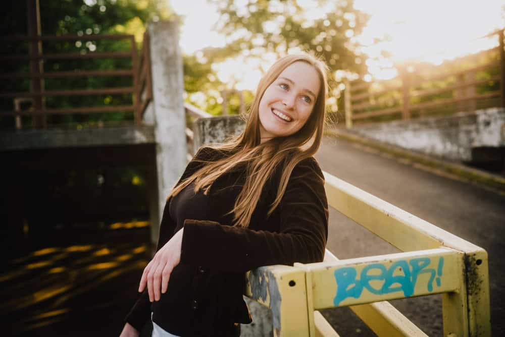 A woman with long auburn hair wearing a dark jacket smiles while leaning on a yellow railing with blue graffiti, radiating self-worth. The background shows a sunlit bridge and lush green trees.
