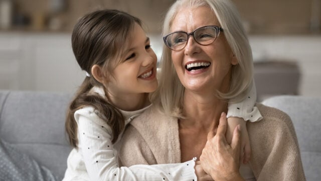 Smiling elderly woman with gray hair and glasses sits on a couch as her grandchild hugs her from behind; both look happy and joyful in a cozy home setting, reflecting the special bond between grandparents and their grandkids.