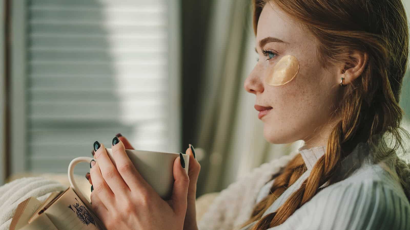 A young woman with braided hair and an under-eye patch holds a white mug of black coffee, gazing thoughtfully out of a window with sunlight streaming in, reflecting on her identity and personal choices.