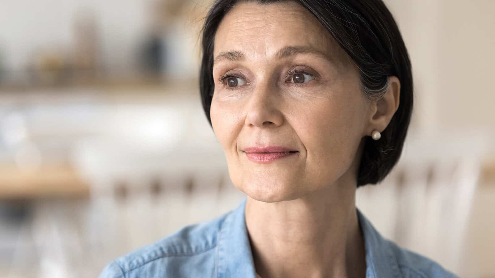 A middle-aged woman with short dark hair and pearl earrings looks thoughtfully into the distance, perhaps reflecting on generational parenting, wearing a light blue shirt with a softly blurred indoor background.