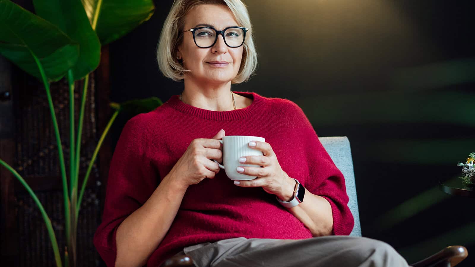 A woman with short blonde hair and glasses, wearing a red sweater and beige pants, sits in a chair holding a white mug, looking at the camera—her calm gaze reflecting her life story of being alone. Green plants and a dark wall are in the background.