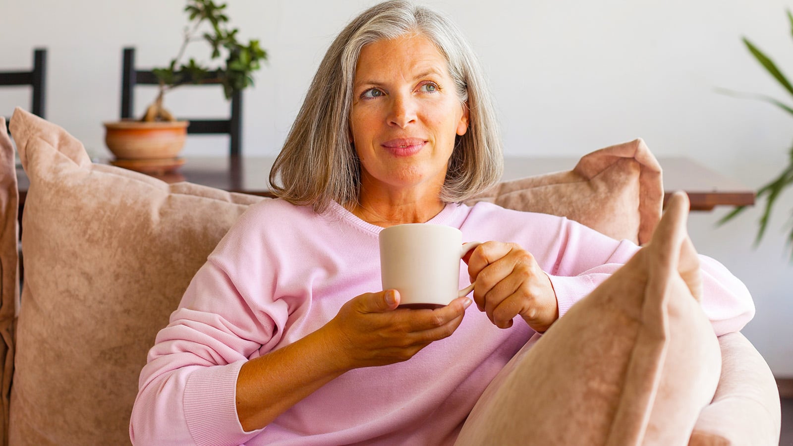 Beautiful middle aged 50 year old female relaxing at home, sitting on comfortable sofa in modern living room interior, looking away and drinking coffee with joyful smile