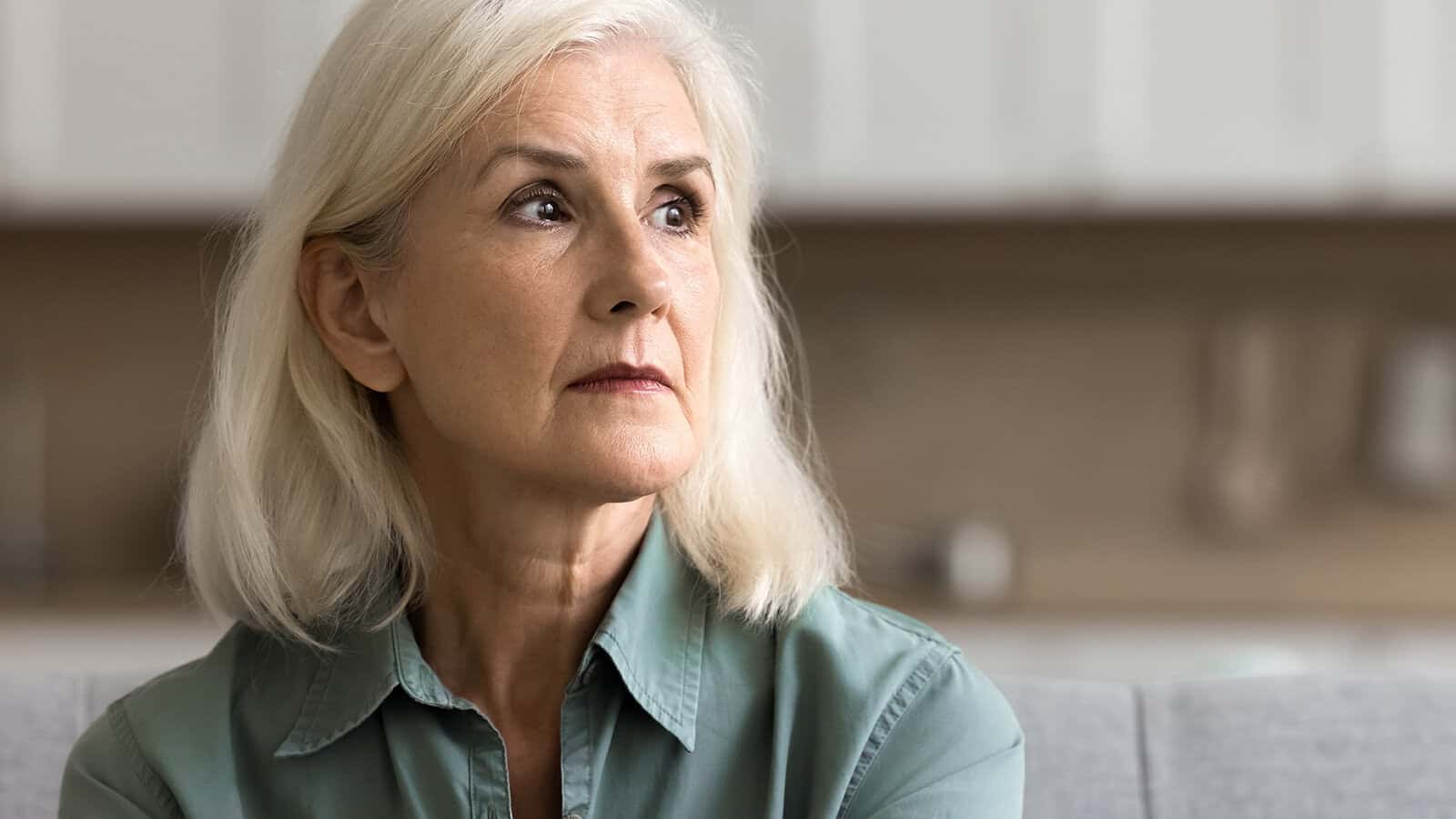 An older woman with white hair and a light green shirt sits indoors, looking thoughtfully to the side with a serious expression—capturing a quiet moment of emotional openness. The background is softly blurred.