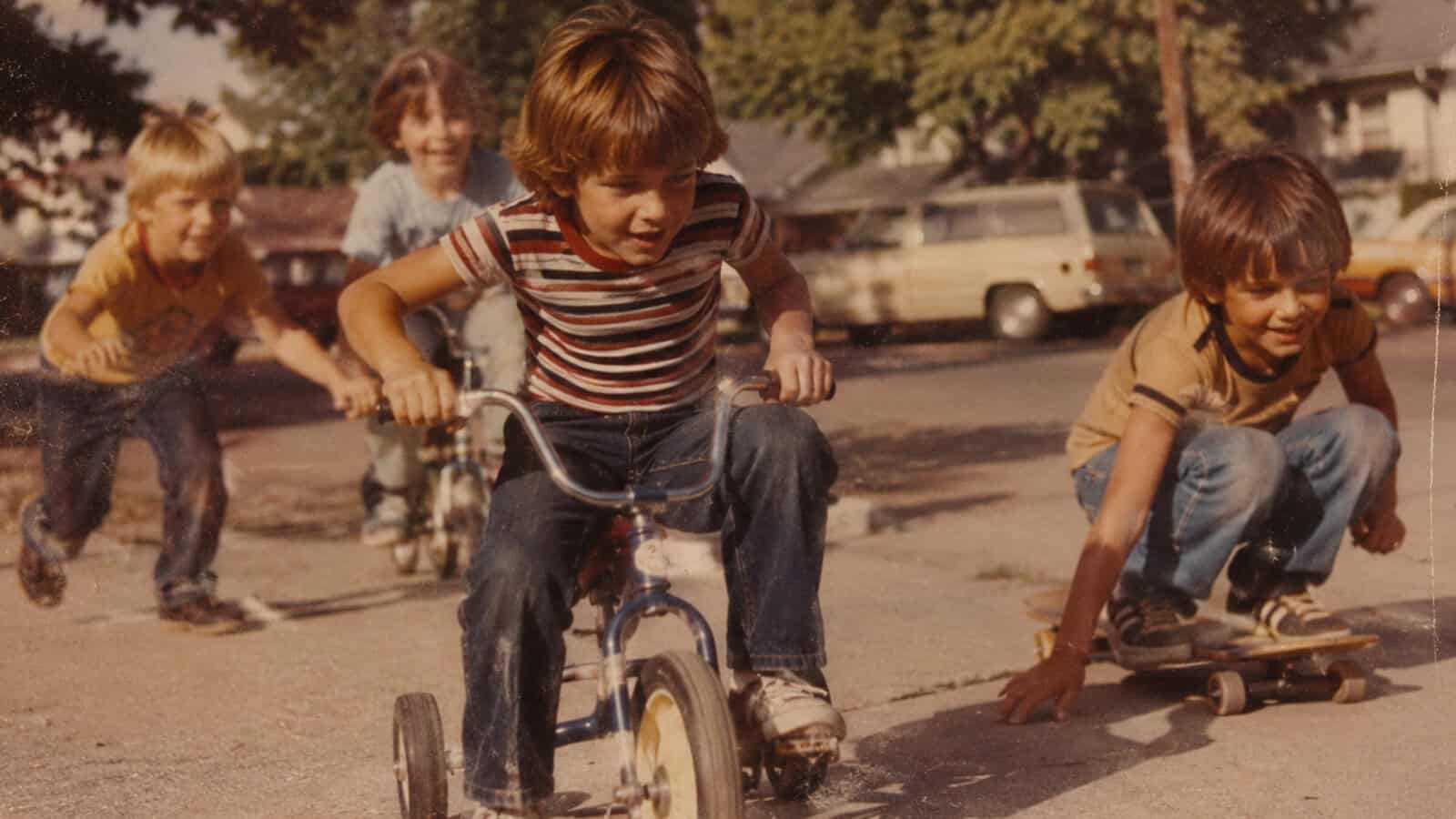 Four children play outside; one rides a tricycle in front, another is on a skateboard, and two run behind. Parked cars and trees set the scene, evoking life lessons and nostalgic memories of the 70s and 80s.