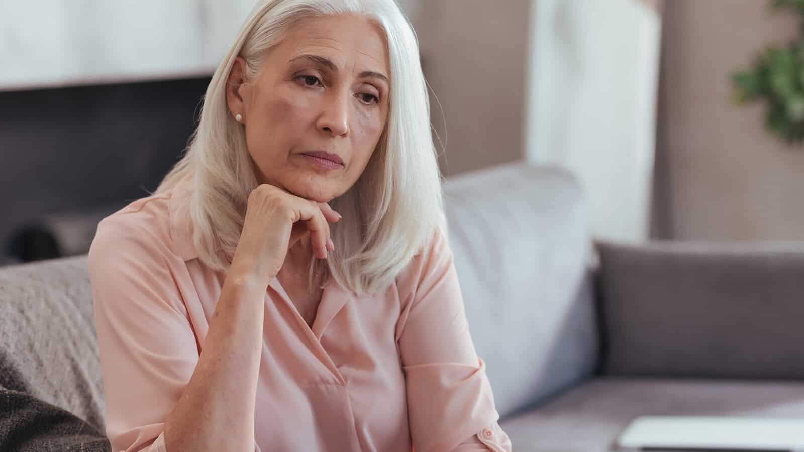 An older woman with long gray hair sits on a couch, resting her chin on her hand and looking thoughtful or concerned—perhaps reflecting on parent child relationships or the psychology after 60. She wears a light pink blouse and appears deep in thought.
