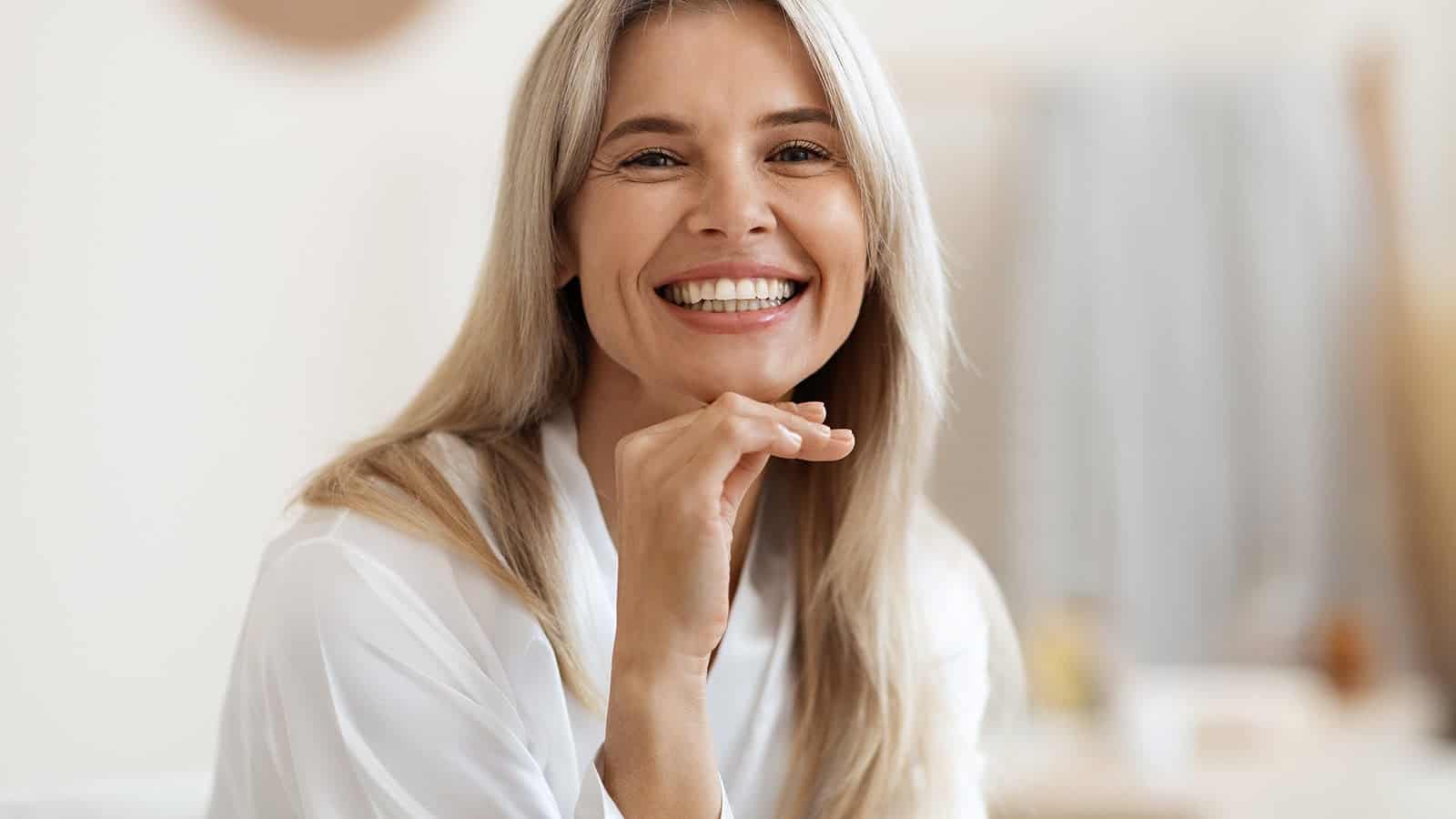 A smiling woman with long blonde hair, feeling like herself, wears a white blouse and rests her chin on her hand. She is indoors, with a soft, neutral background out of focus.