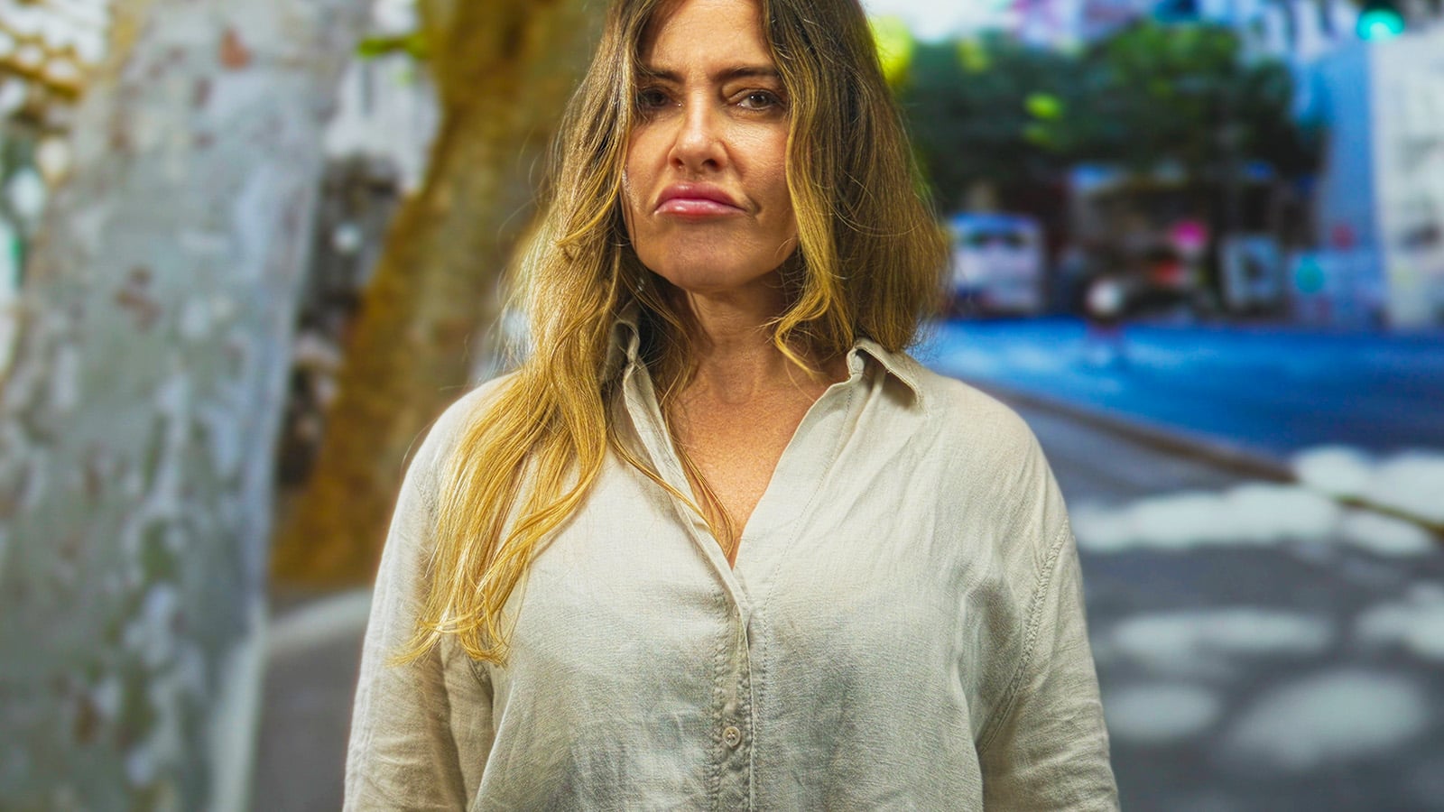 A woman with long, blonde hair wearing a light-colored button-up shirt stands outdoors on a city street, looking directly at the camera with a serious expression. The background is blurred with trees and buildings.