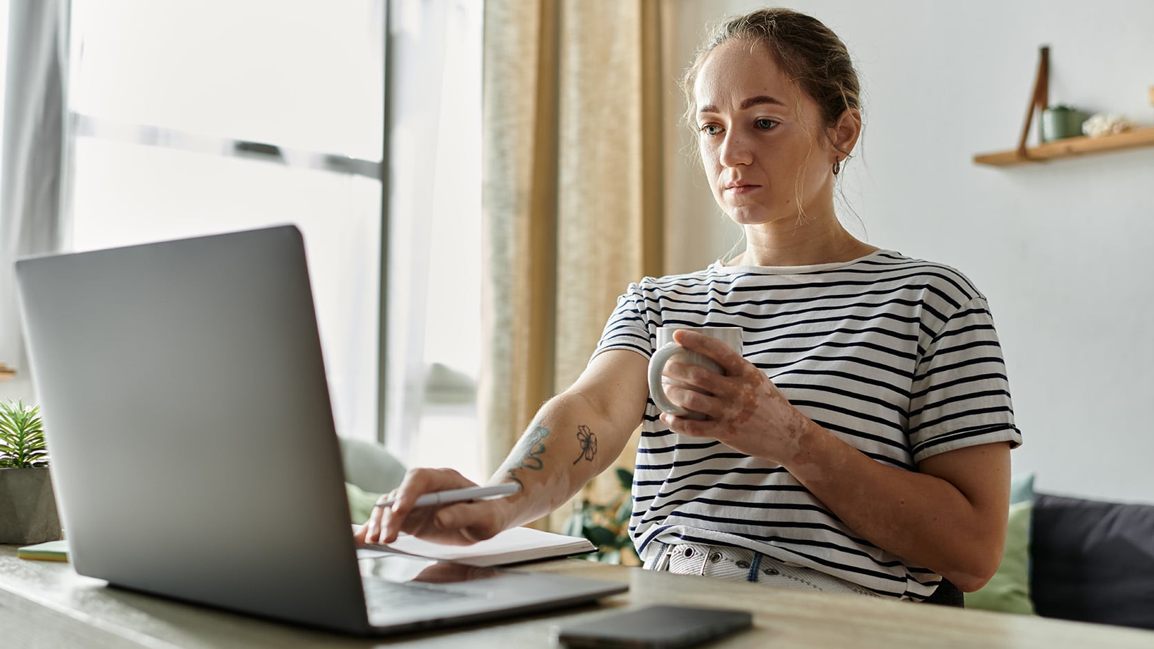 A woman in a striped shirt sits at a table, focused on her laptop with a mug in hand. Surrounded by a notebook, pen, and phone, she reflects the challenges of work-life balance in today’s digital workplace.