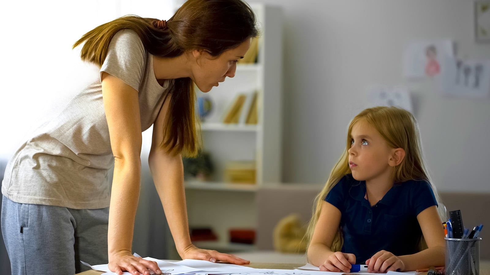 A woman, possibly a psychologist, leans over a table, talking seriously to a young girl with long blonde hair who looks up at her while holding a pen. Papers and pencils are on the table in front of them, and the room is softly lit. The woman's encouraging expression suggests she is urging the girl not to give up and to try harder.