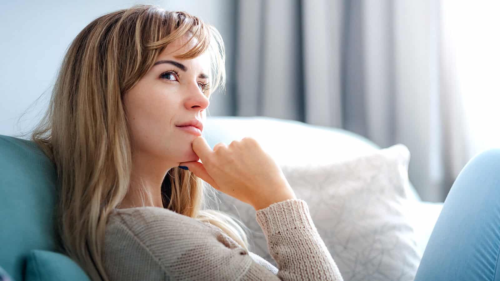 A woman with long, light brown hair sits on a couch, resting her chin on her hand and looking thoughtfully into the distance, as if contemplating letting go of emotional baggage. She wears a beige sweater, with soft daylight filtering through the curtains behind her.