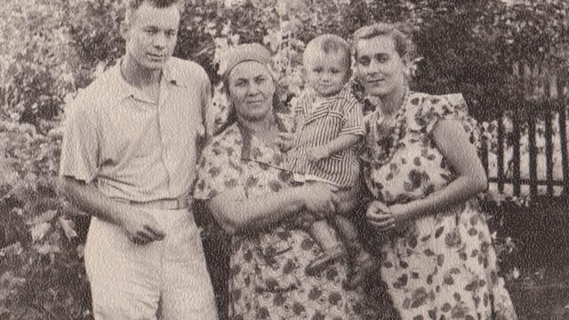 Black-and-white photo of three adults and a toddler outside. One woman holds the child; all wear patterned summer clothes. Vegetation and a wooden fence are visible in the background.