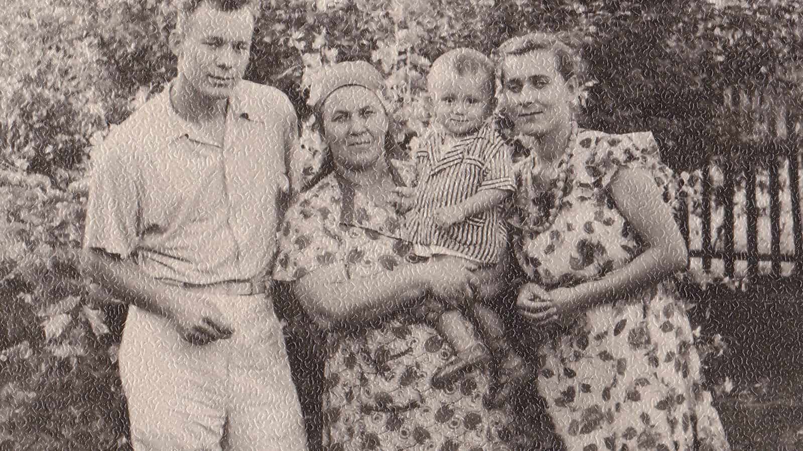 Black-and-white photo of three adults and a toddler outside. One woman holds the child; all wear patterned summer clothes. Vegetation and a wooden fence are visible in the background.