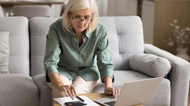 Older woman with gray hair and glasses sits on a sofa, using a calculator and looking at papers on a table in front of her, with an open laptop beside her.