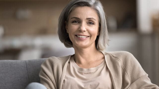 Middle-aged woman with short gray hair, wearing a beige cardigan and cream top, sits on a sofa and smiles at the camera in a softly lit indoor setting.