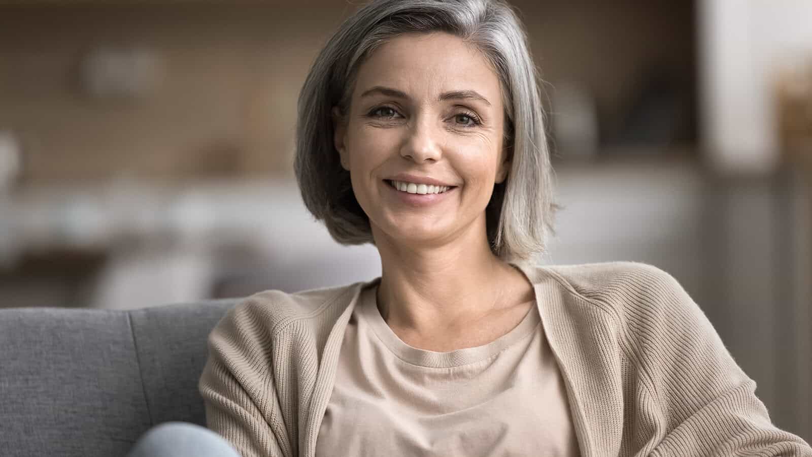 Middle-aged woman with short gray hair, wearing a beige cardigan and cream top, sits on a sofa and smiles at the camera in a softly lit indoor setting.