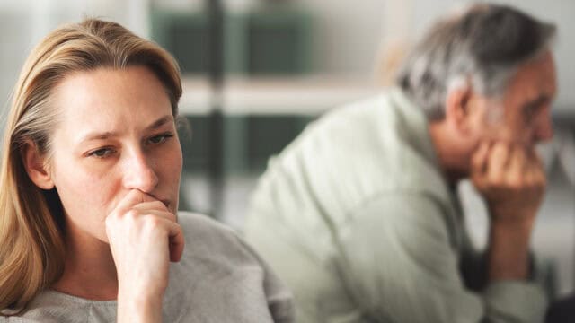 A woman sits in the foreground looking worried with her hand to her mouth, while a man sits in the background facing away, both appearing upset.
