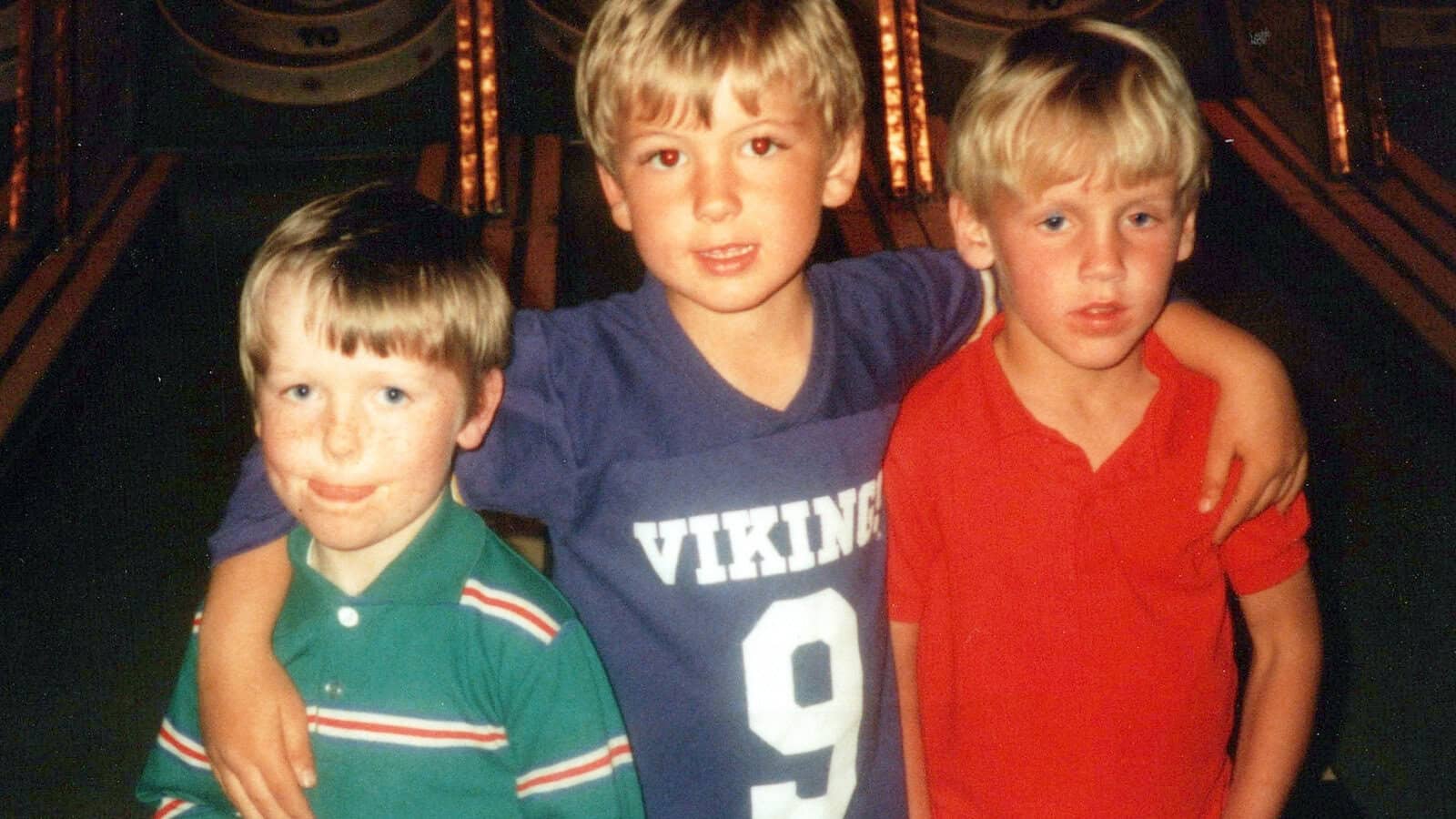 Three young boys stand together indoors. The boy in the center, wearing a purple Vikings 9 shirt, has his arms around the other two boys. The boy on the left wears green stripes; the boy on the right wears a red shirt.