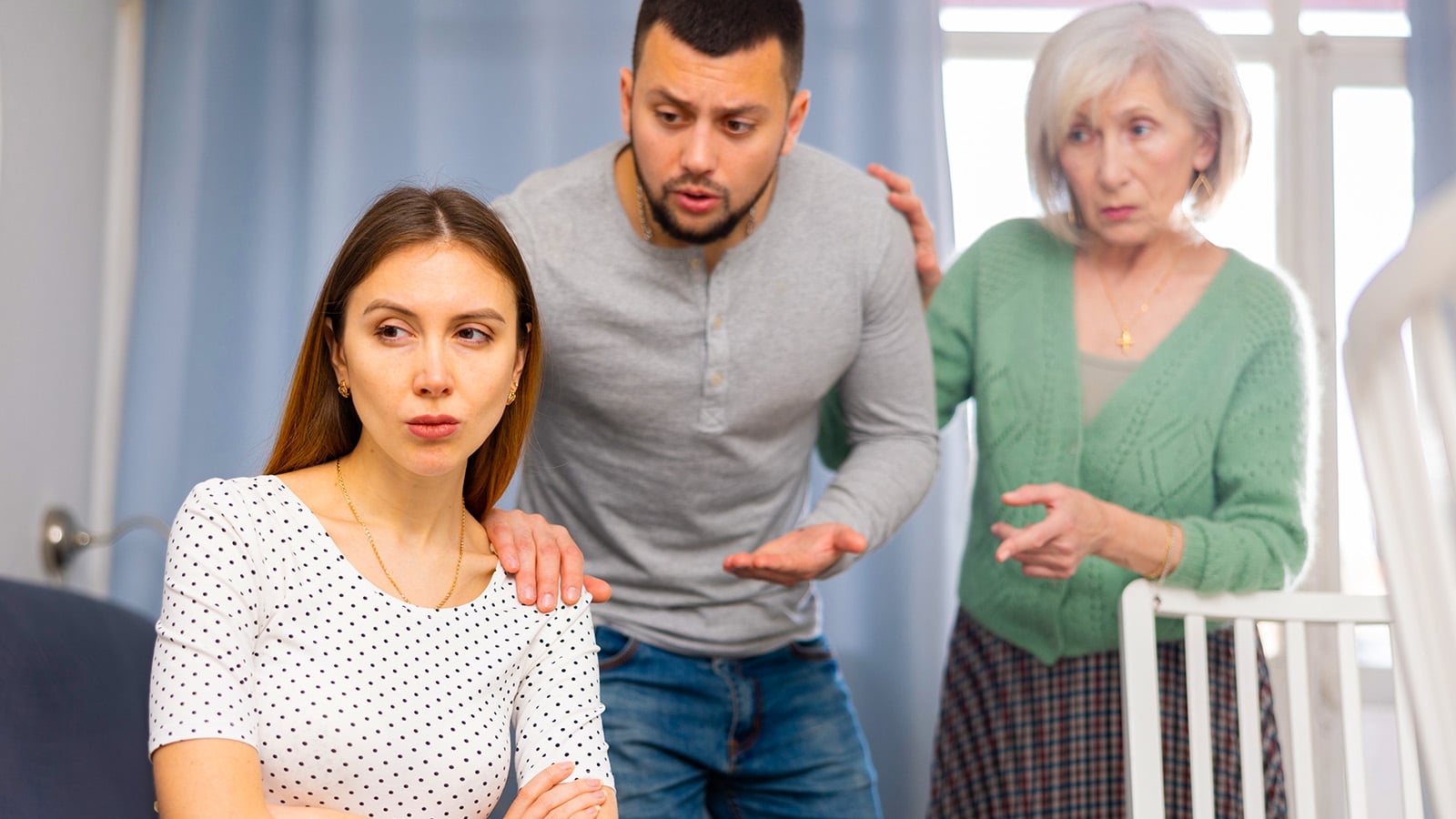 A woman sits with her arms crossed, looking away, while her partner and an older woman—likely her parent—stand behind her, appearing concerned and engaged in a serious conversation about their relationship as an adult child.