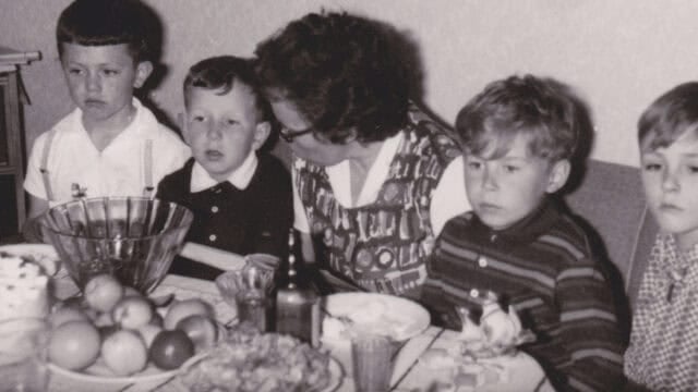 Four children and a woman sit at a table with fruit, drinks, and food. The photo is in black and white, and the group appears to be at a casual gathering or family meal.