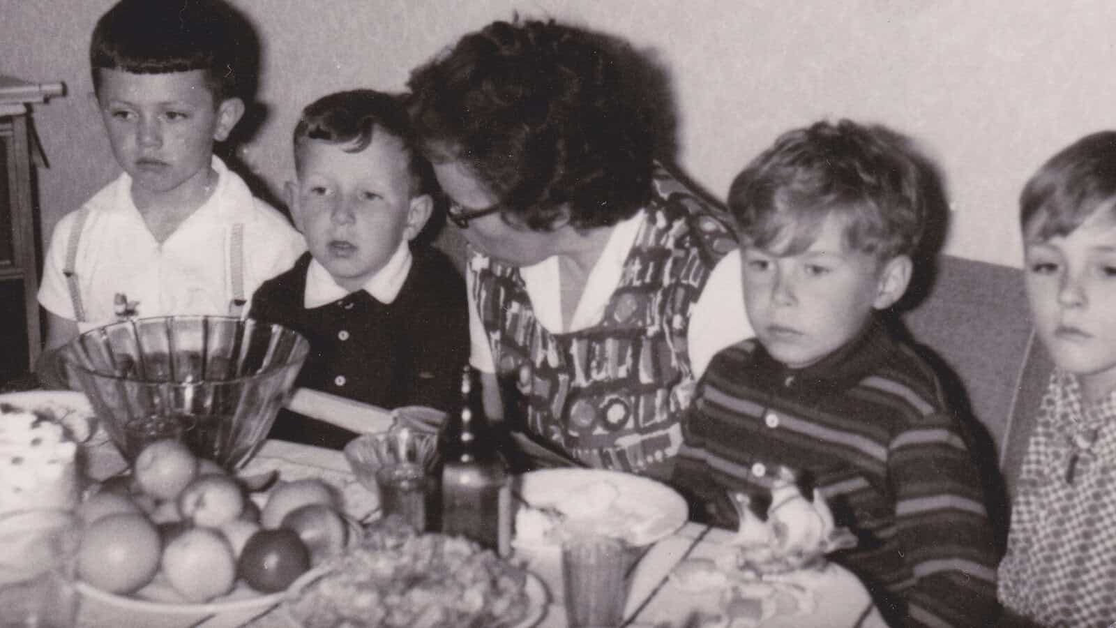 Four children and a woman sit at a table with fruit, drinks, and food. The photo is in black and white, and the group appears to be at a casual gathering or family meal.
