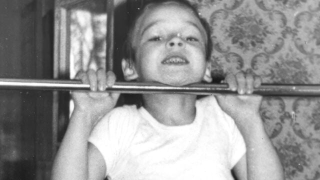 A child in a white shirt smiles while doing a pull-up on a metal bar indoors. The background features patterned wallpaper and a window reflecting trees.