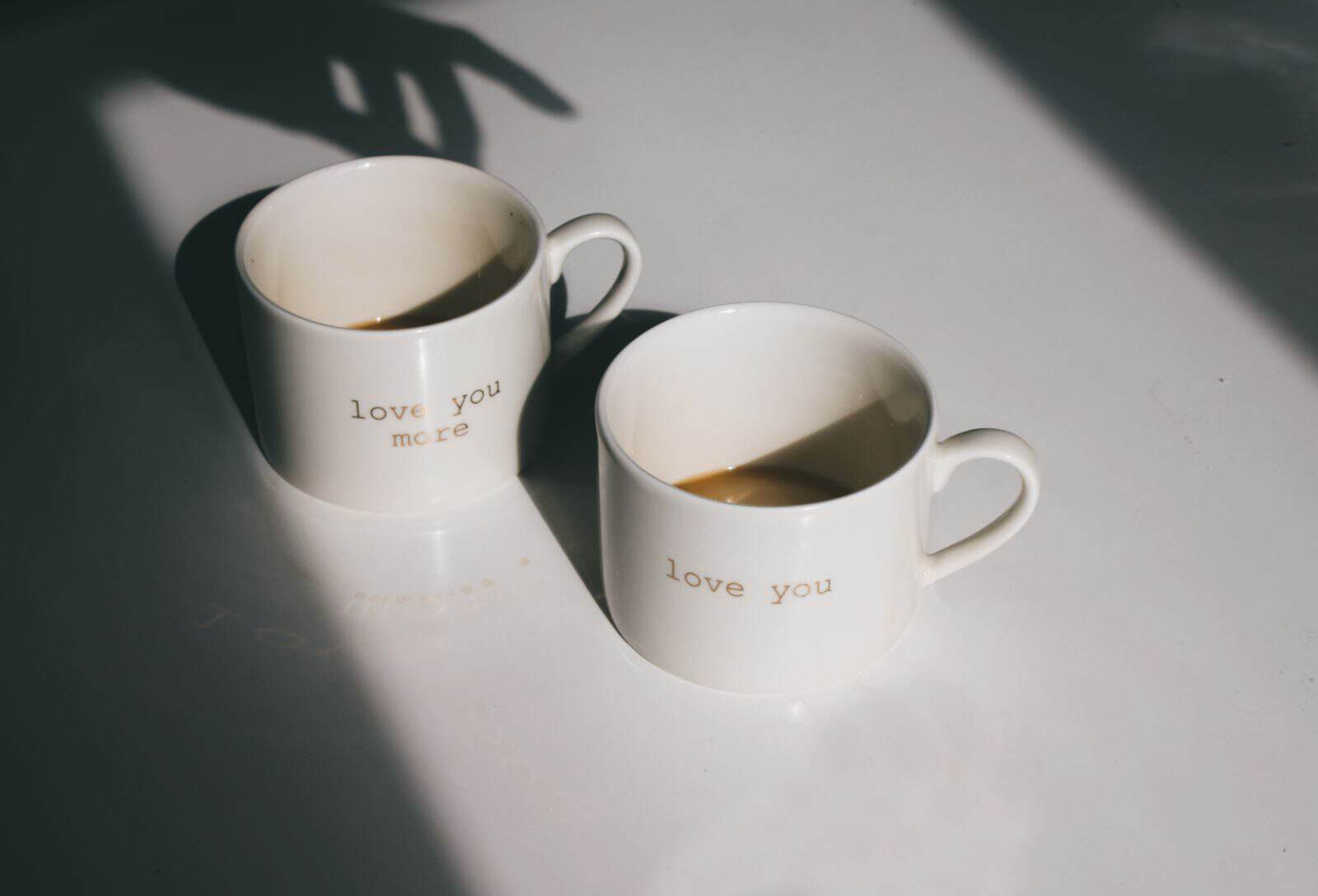 Pair of ceramic mugs with 'love you' text, casting artistic shadows on a white surface.