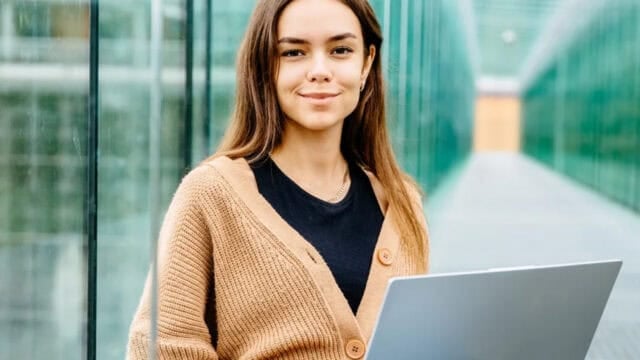 Young woman with long brown hair, wearing a tan cardigan and black shirt, sits outside holding a laptop, with glass walls on both sides and a blurred background.