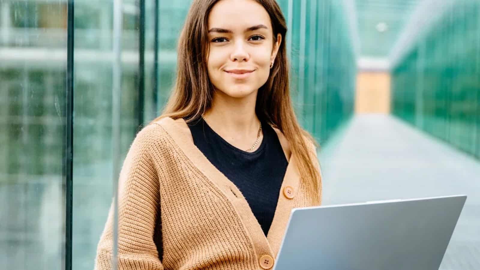 Young woman with long brown hair, wearing a tan cardigan and black shirt, sits outside holding a laptop, with glass walls on both sides and a blurred background.