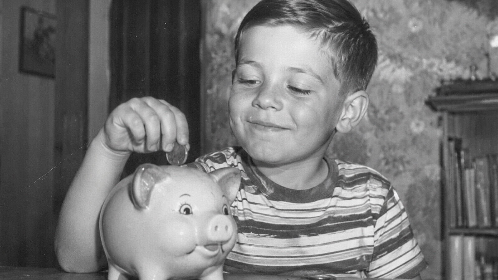 A young boy smiles as he puts a coin into a piggy bank. He is wearing a striped shirt and sitting indoors, with a bookshelf and patterned wallpaper in the background.