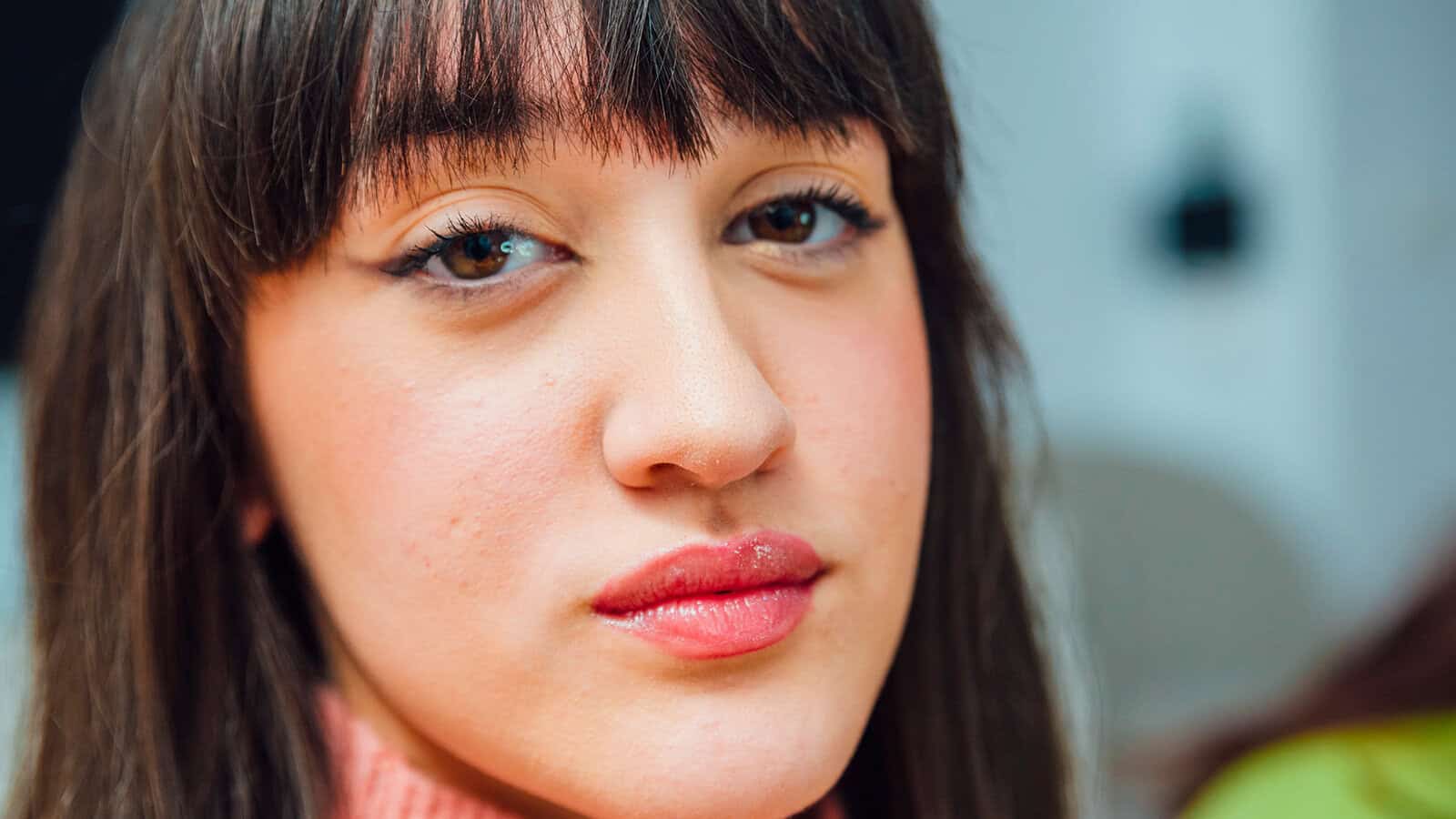 A young woman with straight brown hair and bangs looks into the camera with a neutral expression, wearing a pink sweater. The background is softly blurred.