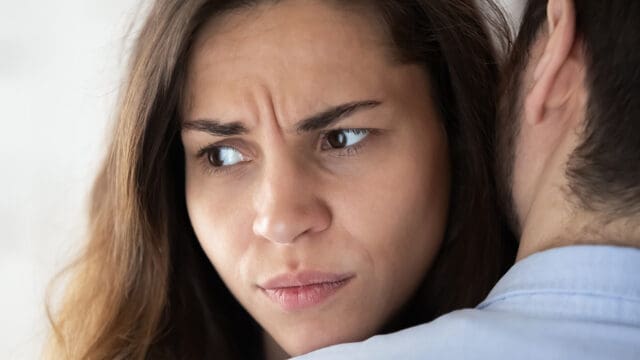 A woman with long brown hair frowns while being hugged by someone, appearing worried or upset as she looks away from the person embracing her.
