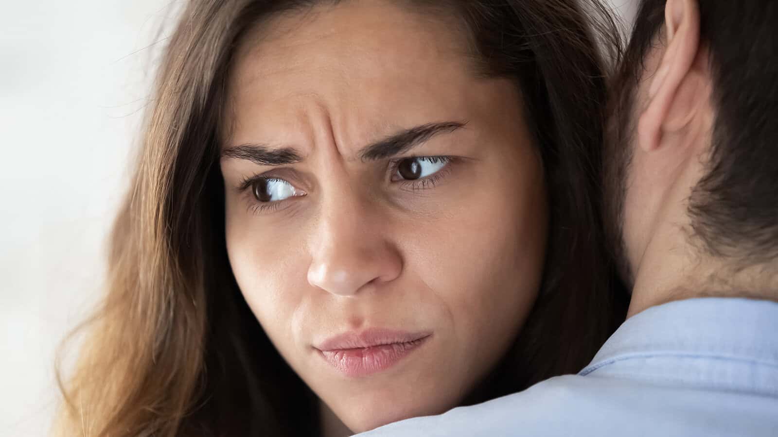 A woman with long brown hair frowns while being hugged by someone, appearing worried or upset as she looks away from the person embracing her.