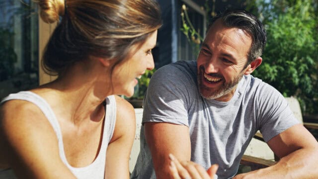 A man and woman sit outdoors, smiling and talking to each other on a sunny day, with greenery and sunlight in the background—a gentle moment that could reflect relationship signs of a peaceful breakup or even a marriage ending on amicable terms.