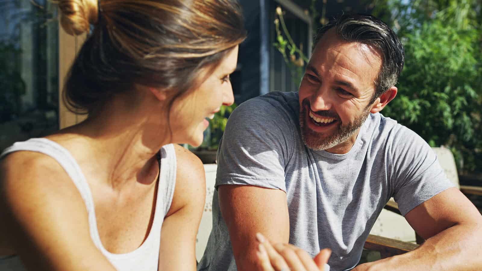 A man and woman sit outdoors, smiling and talking to each other on a sunny day, with greenery and sunlight in the background—a gentle moment that could reflect relationship signs of a peaceful breakup or even a marriage ending on amicable terms.