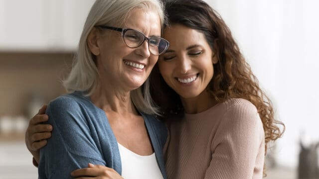 Two women, one older with gray hair and glasses and one younger with long brown hair, smiling and hugging each other warmly indoors, reflect a strong parent-child relationship and clear signs of trust between them.