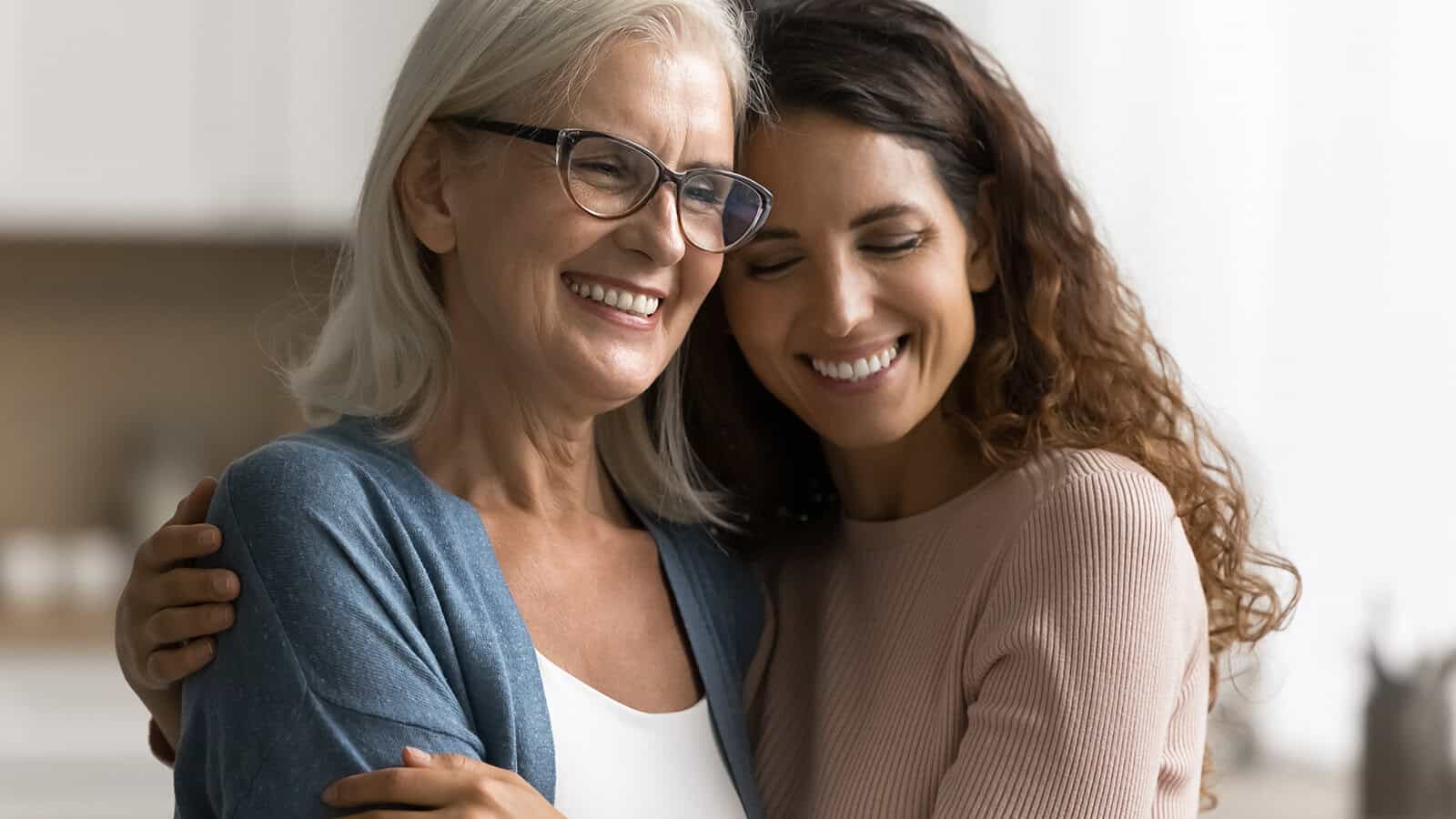 Two women, one older with gray hair and glasses and one younger with long brown hair, smiling and hugging each other warmly indoors, reflect a strong parent-child relationship and clear signs of trust between them.