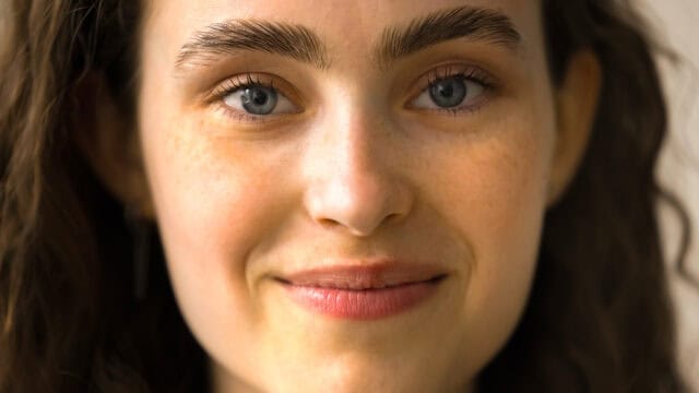 Close-up of a young woman with curly brown hair, blue eyes, full eyebrows, and a slight smile, looking directly at the camera.