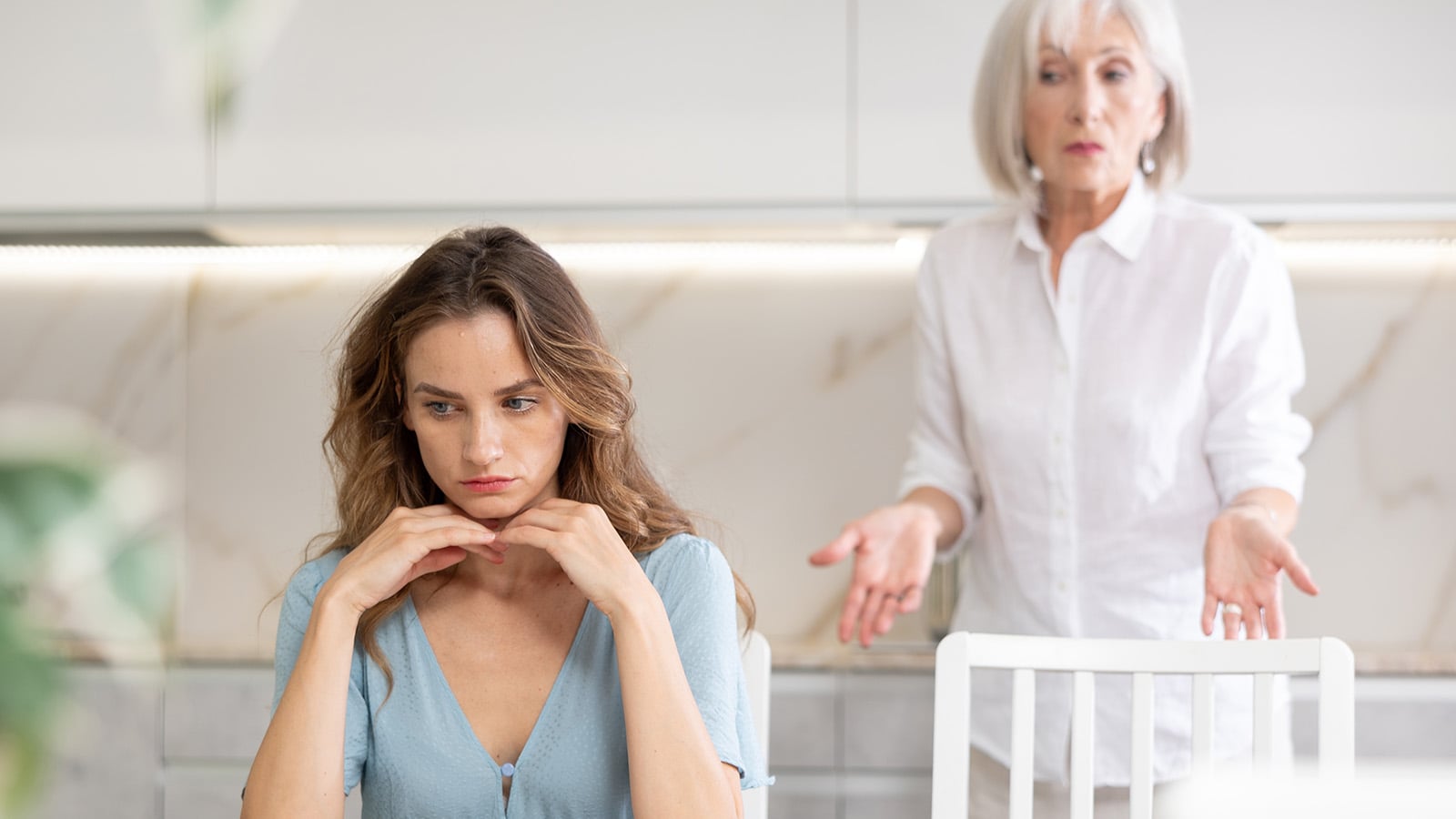 A young woman sits at a table looking upset, while an older woman in the background gestures with her hands, appearing concerned or frustrated—capturing common signs of resentment that can develop from parenting mistakes and affect adult children resentment.