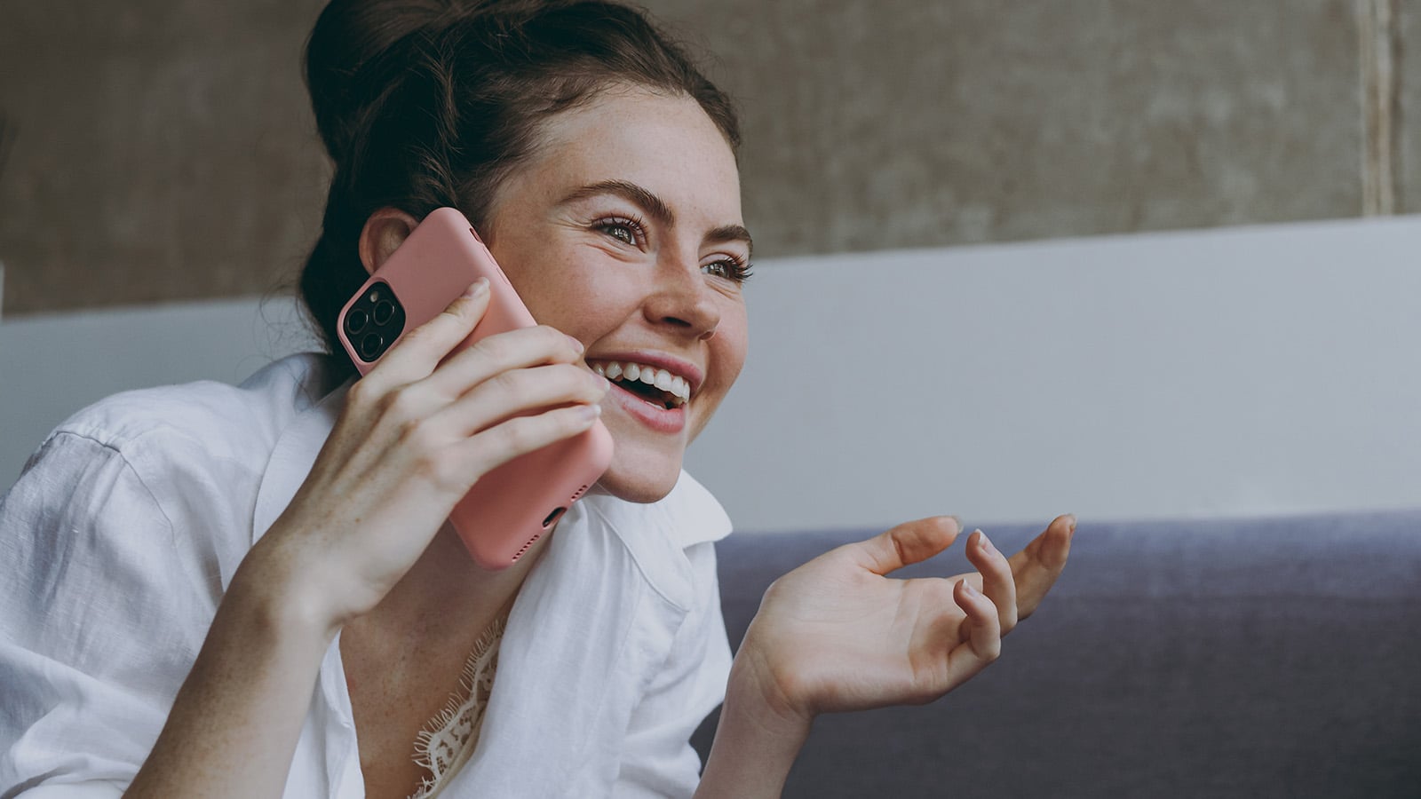 A smiling woman in a white shirt talks on her phone, holding it to her ear with one hand while gesturing with the other, sitting indoors on a sofa. Her warm expression suggests she's nurturing a close relationship, always being kind and attentive during conversations.
